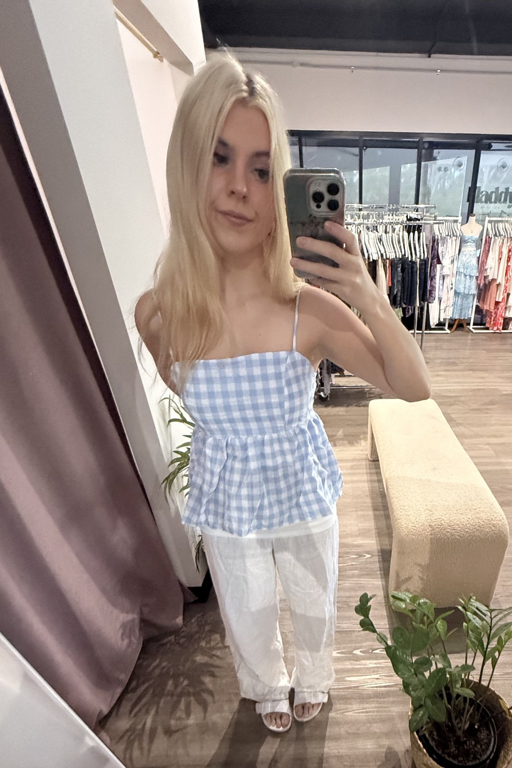 Woman taking a mirror selfie wearing a blue checkered top and white pants in a clothing store.