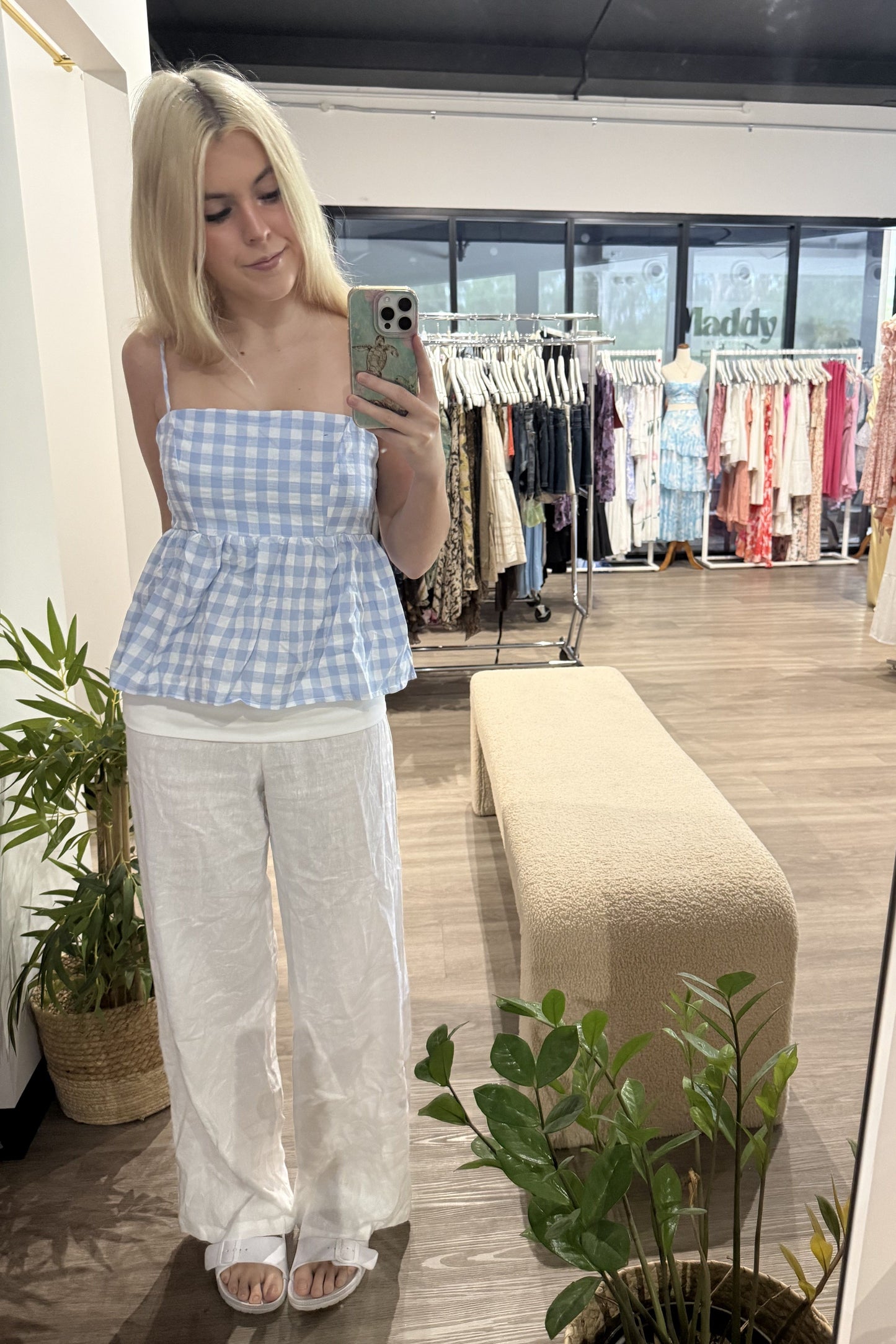 Woman taking a mirror selfie wearing a blue checkered top and white pants in a clothing store.