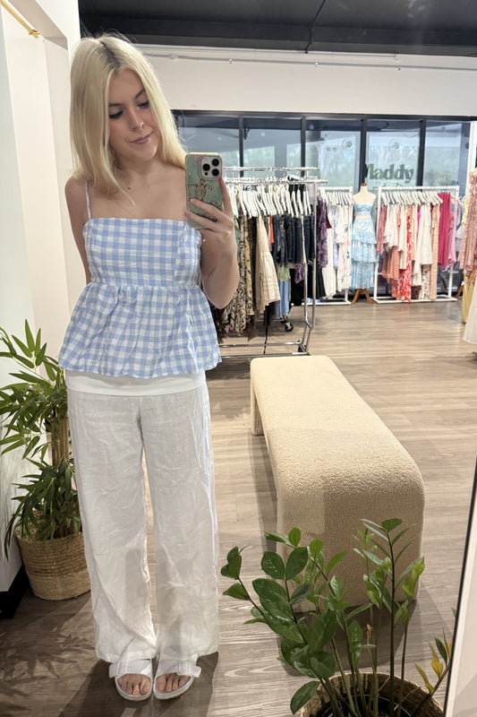 Woman taking a mirror selfie wearing a blue checkered top and white pants in a clothing store.